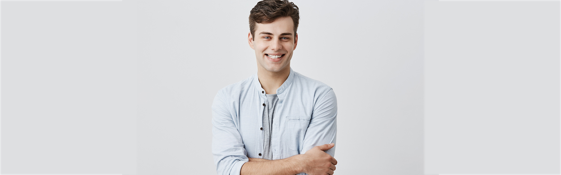 People and lifestyle concept. Portrait of attractive young caucasian male in good mood, wearing blue long sleeved shirt smiling cheerfully showing his perfect white teeth, happy with positive news, keeping his arms folded.
